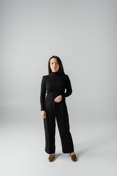 full length of young asian woman in black pants and turtleneck standing on grey  - Foto, immagini