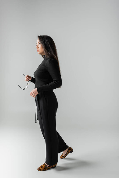 full length of brunette asian woman in black outfit and loafers with animal print holding glasses on grey  - Foto, Imagem