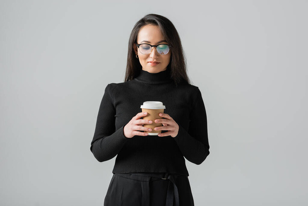 smiling asian woman in stylish black turtleneck and glasses looking at paper cup isolated on grey  - Фото, зображення