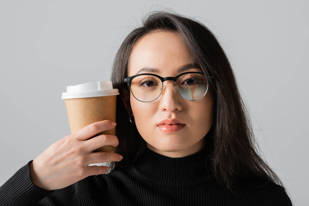 brunette asian woman in stylish black turtleneck and glasses holding coffee to go isolated on grey  - Foto, imagen