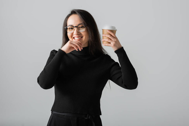 happy asian woman in black turtleneck and glasses holding coffee to go isolated on grey  - 写真・画像