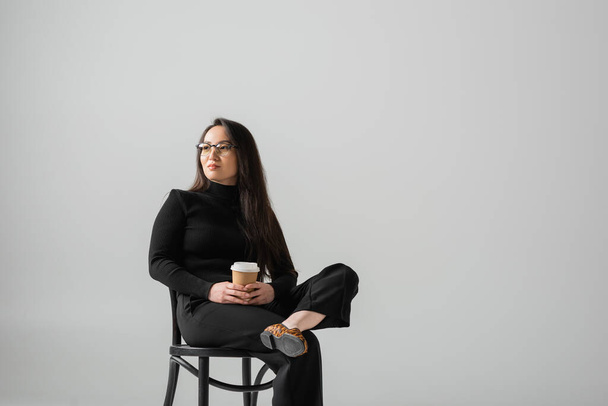 brunette asian woman in black outfit and glasses holding paper cup while sitting on chair isolated on grey  - Foto, afbeelding