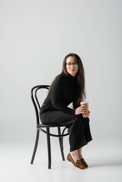 full length of brunette asian woman in black outfit and glasses holding paper cup while sitting on chair on grey  - Fotó, kép