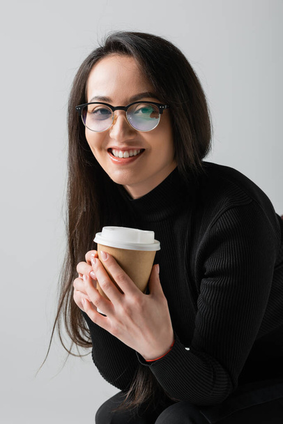 joyful asian woman in black turtleneck and glasses holding paper cup isolated on grey  - Photo, Image