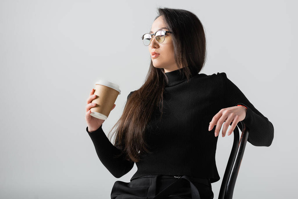 asian woman in black turtleneck and glasses holding paper cup while sitting on chair isolated on grey  - Фото, изображение