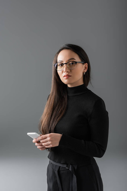 brunette asian woman in black turtleneck and glasses holding smartphone on dark grey  - Fotó, kép