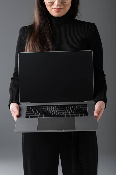 обрізаний вид усміхненої жінки в чорному черепашку, що показує ноутбук з порожнім екраном ізольовано на темно-сірому
  - Фото, зображення
