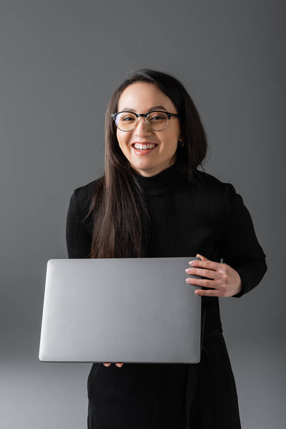 joyful asian woman in black turtleneck and glasses holding laptop on dark grey  - Foto, Imagem