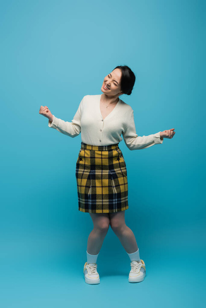 full length of joyful asian woman in checkered skirt and white sneakers standing on blue  - 写真・画像