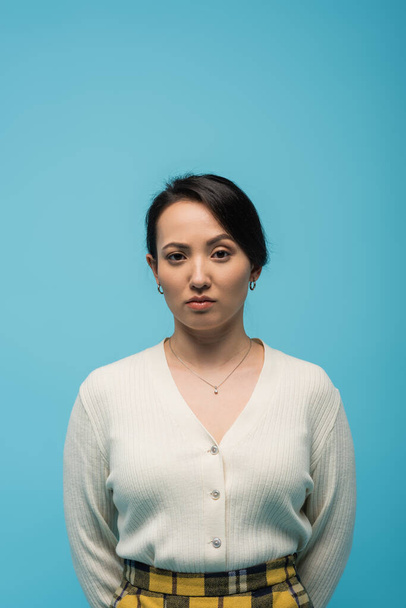 Серйозна азіатська жінка в кардигані дивиться на камеру ізольовано на синьому
  - Фото, зображення