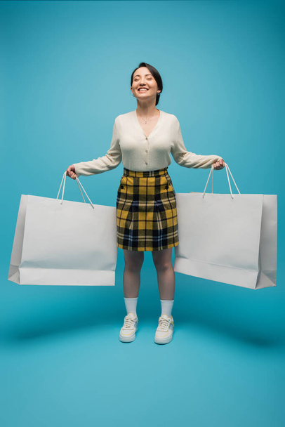 Positive asian woman holding shopping bags while standing on blue background - Foto, afbeelding