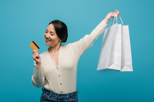 Cheerful asian woman holding credit card and shopping bags isolated on blue  - Foto, Imagem