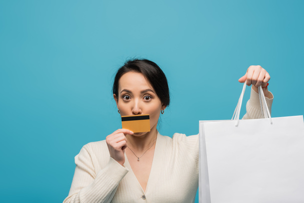 Young asian woman holding credit card near face and shopping bags isolated on blue  - Foto, Imagen
