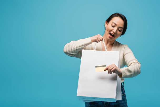 Excited asian customer holding shopping bags and credit card isolated on blue  - Photo, Image