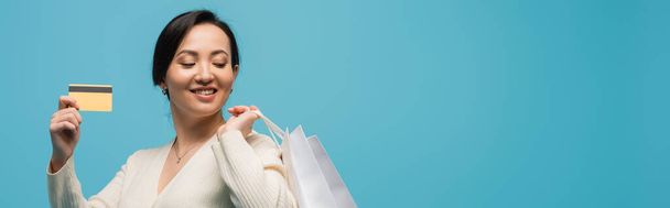 Positive asian shopaholic holding shopping bags and credit card isolated on blue, banner  - Foto, Imagen