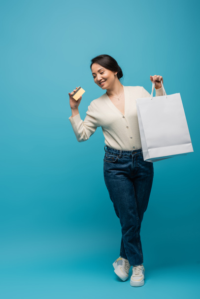 Cheerful asian woman in cardigan and jeans holding shopping bags and credit card on blue background - Φωτογραφία, εικόνα