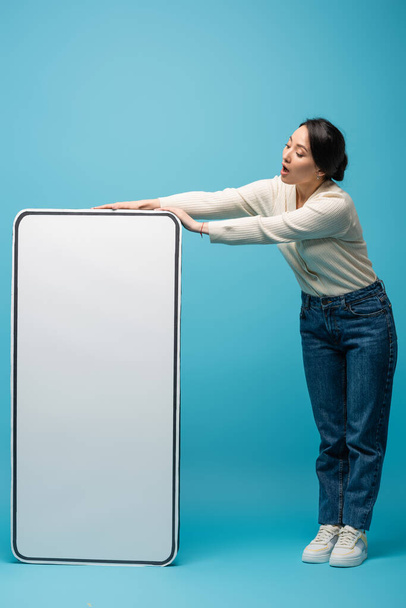 Shocked asian woman in jeans and cardigan standing near big smartphone model on blue background - Фото, изображение