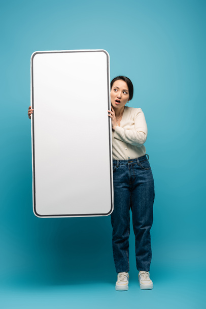 Shocked asian woman holding big smartphone model on blue background - Photo, Image