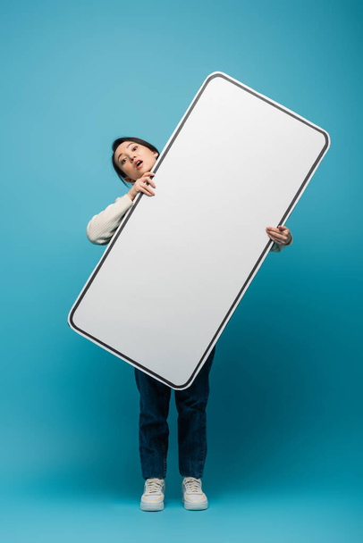 Confused asian woman holding big smartphone model on blue background - Фото, изображение