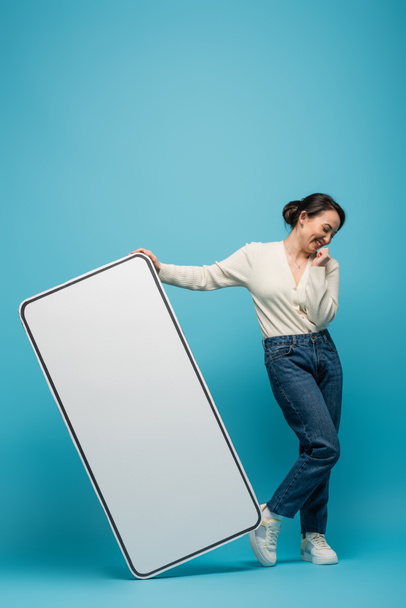 Cheerful asian woman standing near big smartphone mockup on blue background - Photo, Image