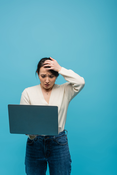 Stressed asian freelancer holding laptop and touching head isolated on blue  - Photo, image