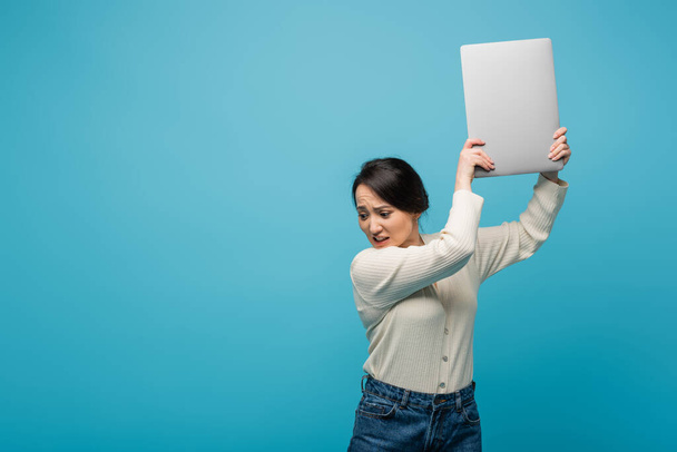 Angry asian woman holding laptop above head isolated on blue  - Photo, image