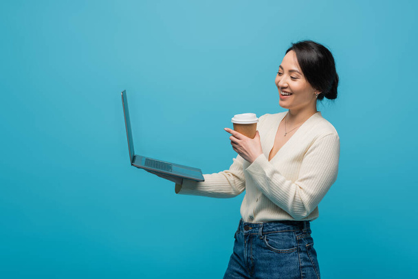 Позитивна азіатська жінка тримає паперову чашку і ноутбук ізольовано на синьому
  - Фото, зображення