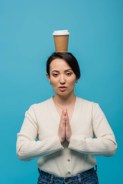 Asian woman with coffee to go on head doing praying hands gesture isolated on blue  - Fotó, kép