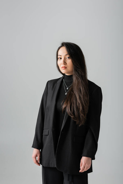 Young asian woman in suit and turtleneck looking at camera isolated on grey  - Zdjęcie, obraz