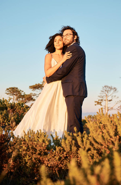 Interracial couple, love and wedding at sunset, hug and happy for romance and relationship in nature. Marriage, bride and groom hug at outdoor ceremony and sweet on blue sky background space. - Photo, Image