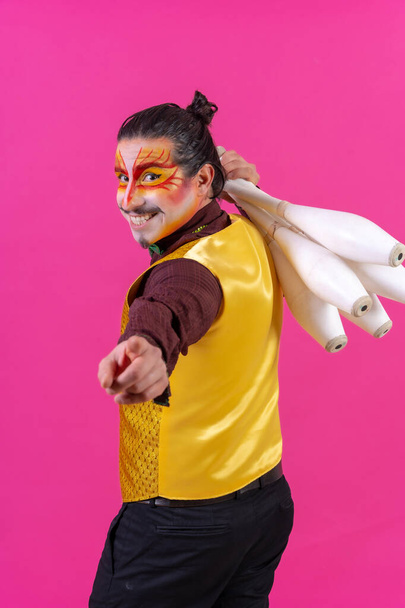Juggler in a vest and with painted face juggling maces on a pink background, pointing - Foto, imagen