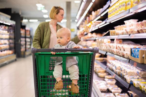 Mother pushing shopping cart with her infant baby boy child down department aisle in supermarket grocery store. Shopping with kids concept - Foto, immagini