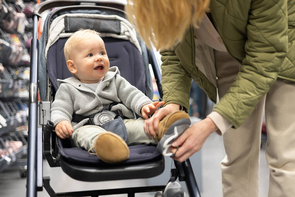 Casualy dressed mother choosing sporty shoes and clothes products in sports department of supermarket store with her infant baby boy child in stroller - Foto, Bild