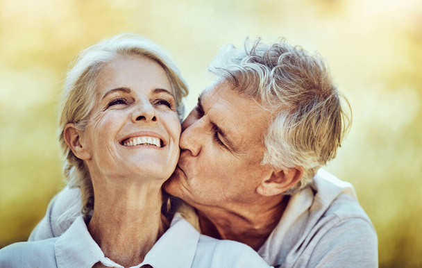Senior couple, kiss and cheek with smile for love, romance and embrace together in the nature outdoors. Happy elderly woman and man kissing face for romantic bonding or quality time in happiness. - Photo, image