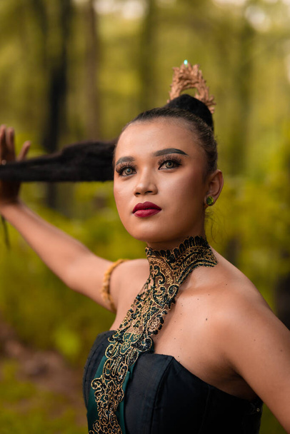 Gorgeous Asian woman in makeup with black hair and red lipstick wearing golden jewelry during the dance performance in front of the jungle - Fotoğraf, Görsel