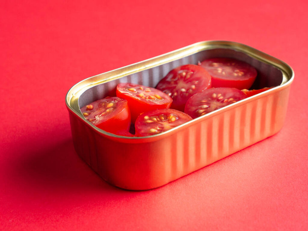 close-up blik van een blik met enkele kerstomaten gesneden in helften, geïsoleerd op rode achtergrond, voeding en conserven voedsel concept - Foto, afbeelding