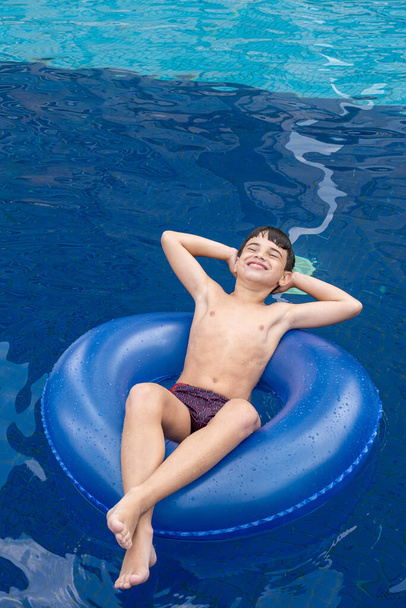 Smiling Brazilian 9 year old in the pool on a sunny day, with arms outstretched and sitting on a buoy. - Photo, image