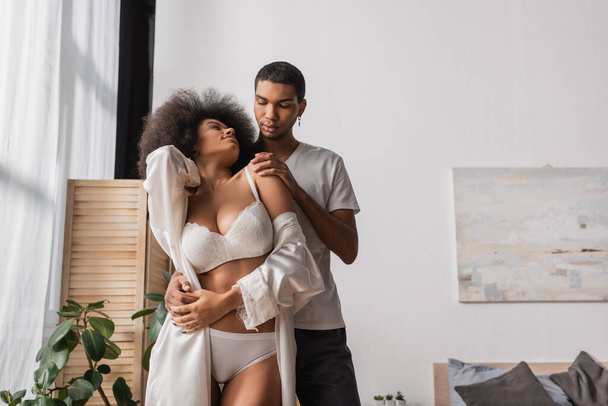 jeune homme afro-américain embrassant petite amie bouclée portant une lingerie sexy dans la chambre - Photo, image