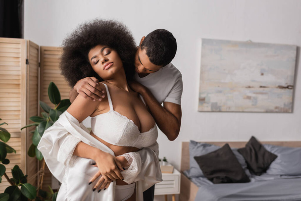 афроамериканський чоловік обіймає спокусливу дівчину в білому бюстгальтері, що стоїть із закритими очима в спальні
 - Фото, зображення