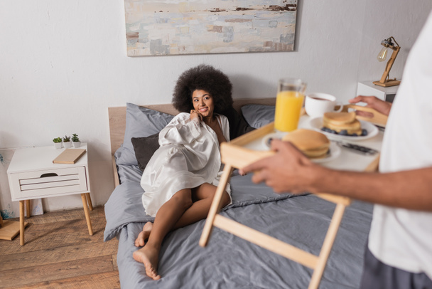 seductora mujer afroamericana en bata de seda acostada en la cama cerca de un hombre borroso sosteniendo bandeja con desayuno - Foto, Imagen