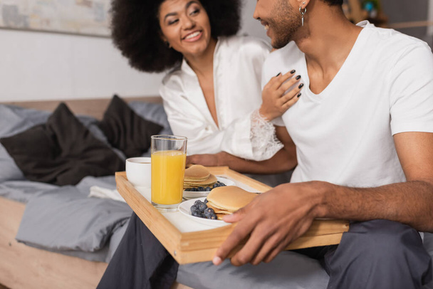 lachende Afrikaans-Amerikaanse vrouw aanraken vriendje houden lade met heerlijke pannenkoeken en sinaasappelsap in de slaapkamer - Foto, afbeelding