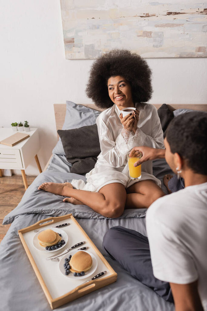 сексуальная африканская американка в шелковом халате сидит на кровати с апельсиновым соком рядом с блинами с ягодами и бойфрендом с чашкой кофе - Фото, изображение