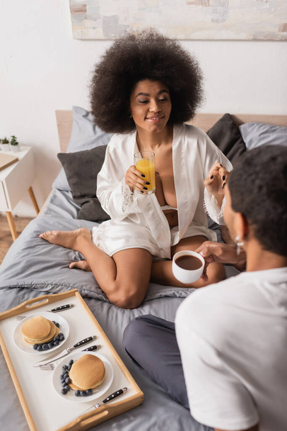 verleidelijk afrikaanse vrouw in wit gewaad met sinaasappelsap in de buurt van vriendje met koffiekopje en pannenkoeken op bed - Foto, afbeelding
