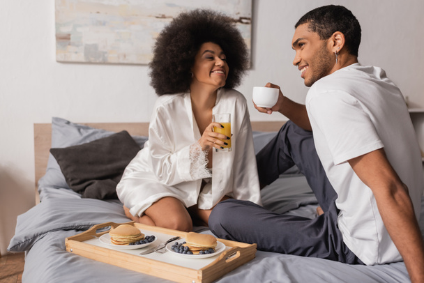 feliz pareja afroamericana sentada con café y jugo de naranja cerca de bandeja con panqueques en el dormitorio - Foto, imagen