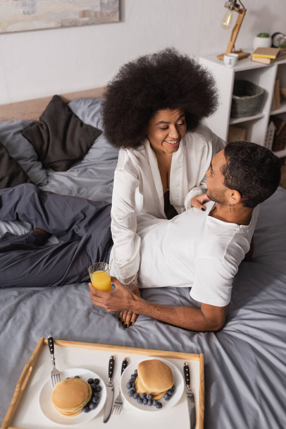 vista de ángulo alto de la mujer afroamericana sonriendo cerca de novio joven y deliciosos panqueques con arándanos en la cama - Foto, Imagen