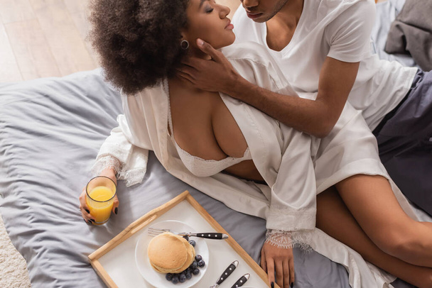 vista de ángulo alto de hombre afroamericano tocando novia en lencería sexy cerca de jugo de naranja y panqueques en la cama - Foto, imagen