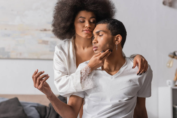sexy african american woman with curly hair and closed eyes touching face of young boyfriend in bedroom - Foto, Imagen