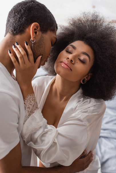 mujer afroamericana caliente en túnica de seda blanca abrazándose con novio joven en el dormitorio - Foto, imagen