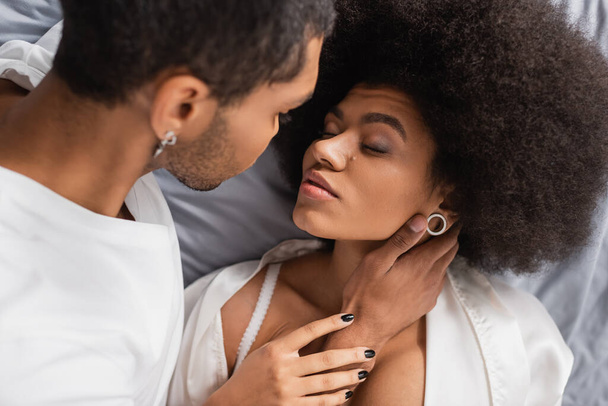 high angle view of african american man near seductive girlfriend lying on bed with closed eyes - Photo, Image
