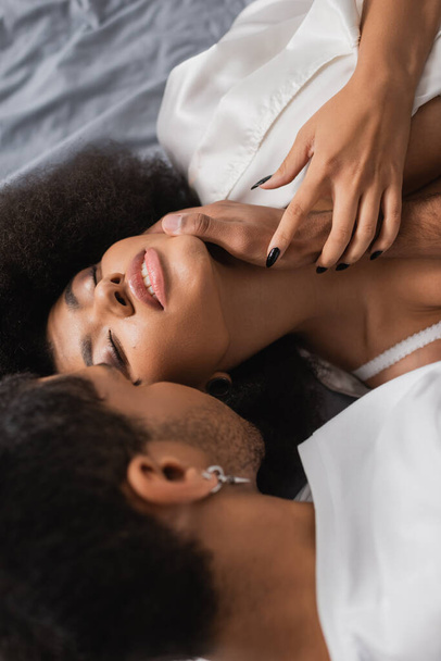 θέα υψηλής γωνίας του νεαρού άνδρα που αγγίζει σέξι αφροαμερικανή φίλη ξαπλωμένη στο κρεβάτι με κλειστά μάτια - Φωτογραφία, εικόνα